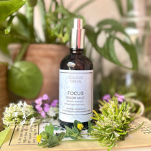 Amber glass bottle of College Green Focus room mist with silver atomiser, sitting on a book surrounded by plants and flowers