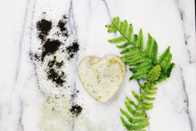 An unwrapped heart shaped bar of Peppermint and Rosemary soap on a white marble surface with coffee grounds and green fern leaves