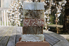 A stack of five unwrapped soaps on a wooden soap dish. Tree in blossom in the background
