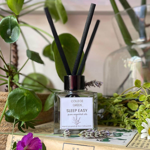 A heavy glass jar of College Green Sleep Easy reed diffuser with three extra thick black reeds on a bedside cabinet surrounded with flowers and greenery