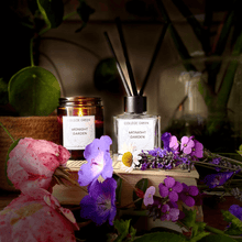 College Green Midnight Garden soy wax candle in amber glass jar and reed diffuser in heavy clear glass jar. Sitting on a pile of books surrounded with summer garden flowers including rose and geranium