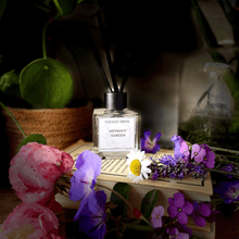 College Green Midnight Garden reed diffuser in heavy clear glass jar. Sitting on a pile of books surrounded with summer garden flowers including rose and geranium
