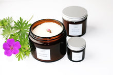 three amber glass jars of Rosy Posy bath soak on a white background with purple geranium flower. One jar is open, showing the contents