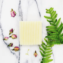 A light coloured bar of unwrapped soap on a marble base next to dried rosebuds and green leaves.