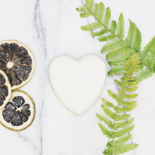 Unwrapped heart shaped bar of soap on marble surface next to slices of lime and green fern leaves
