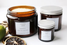 Three amber glass jars of Happy Bath Soak, one open to reveal the contents, two with silver lids,  a lime and lime slices