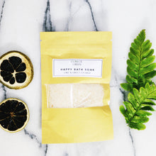 A paper sachet of Happy Bath Soak on a marble work top next to slices of lime and green fern leaves