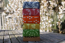 A stack of five wrapped soaps standing on a wooden soap dish. Tree with spring blossom in the background